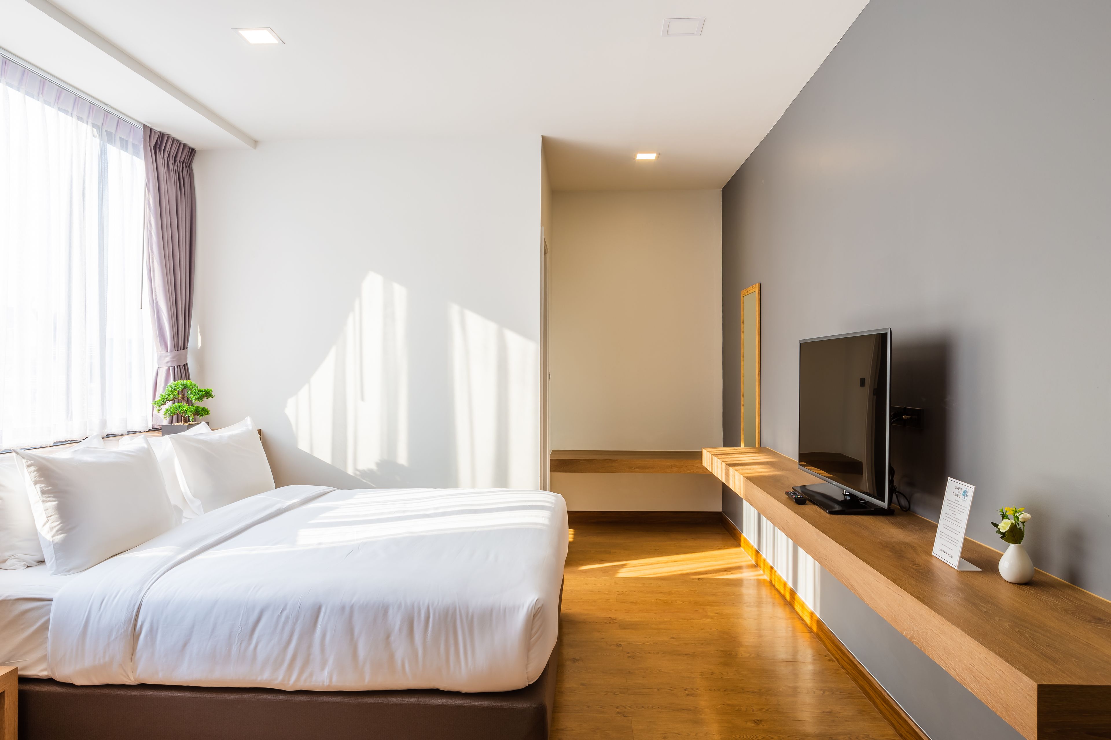 Una habitación de hotel moderna con una cama grande, una mesita de noche de madera con un televisor y vistas al pasillo.
