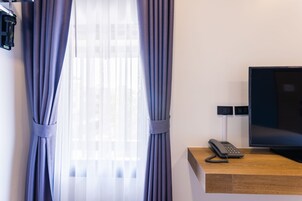 Ett hotellrum med ett skrivbord i trä, en platt-TV, en telefon och ett fönster med tunna, genomskinliga gardiner.