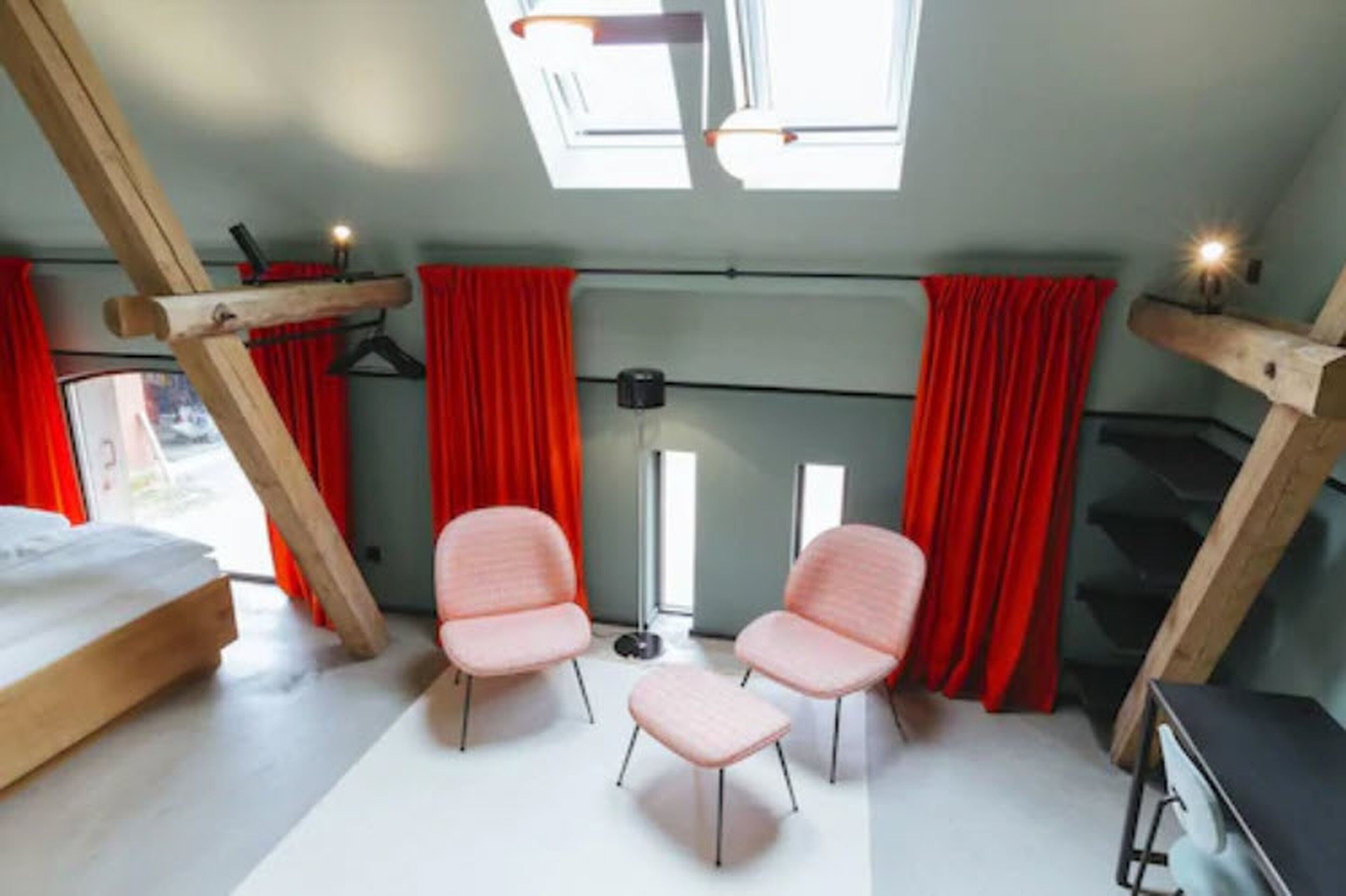 Une pièce avec un plafond en pente, deux fauteuils roses, un lit et des rideaux rouges.