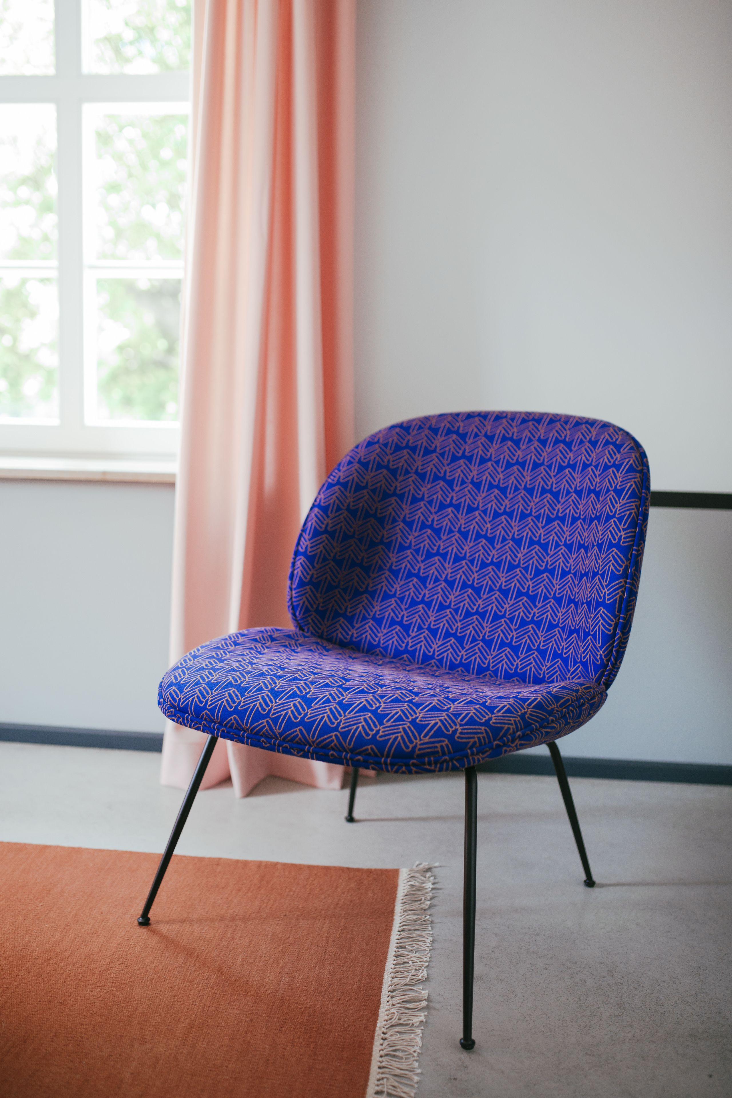 A blue patterned chair with black legs in a room with a window, a pink curtain, and a brown rug.