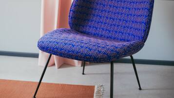 A blue patterned chair with black legs in a room with a window, a pink curtain, and a brown rug.