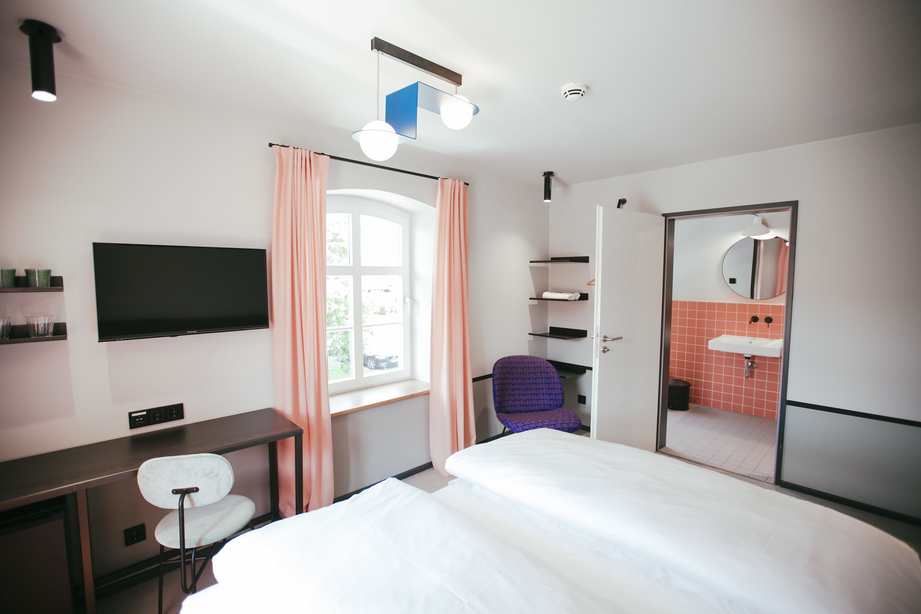 A hotel room with a bed, a desk with a chair, a television, a window with curtains, a bathroom with a mirror and sink, and a door.