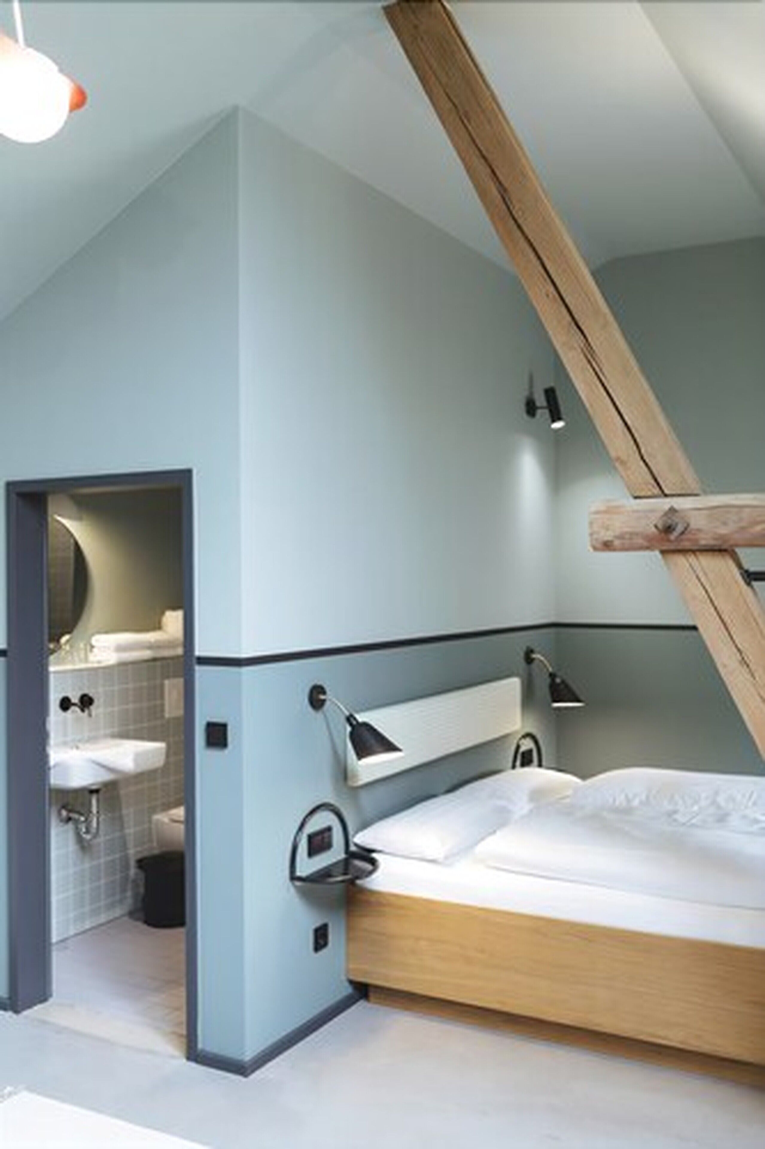 A bedroom with a sloped ceiling, a wooden beam, a bed, a bedside table, a mirror, and a bathroom visible through an open door.