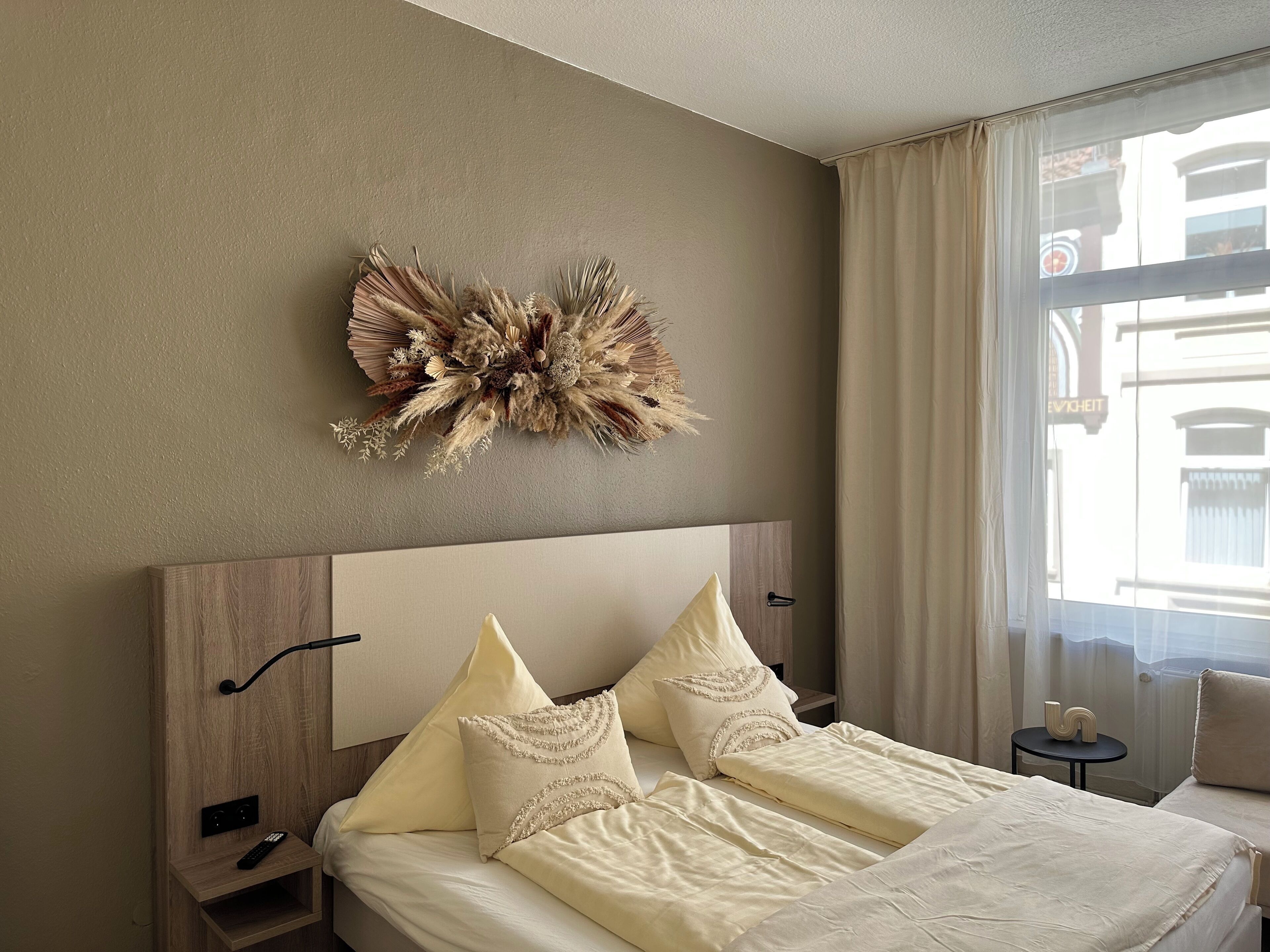 A hotel room with a large bed, a headboard, a wall-mounted decorative piece, a nightstand, and a window with curtains.