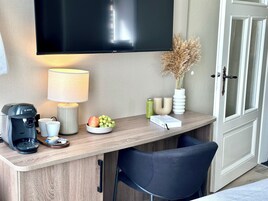 Ein Hotelzimmer mit einem Holztisch, einem Flachbildfernseher, einer Lampe, einer Kaffeemaschine, einer Obstschale und einer Vase mit Trockenblumen sowie einem Sessel.