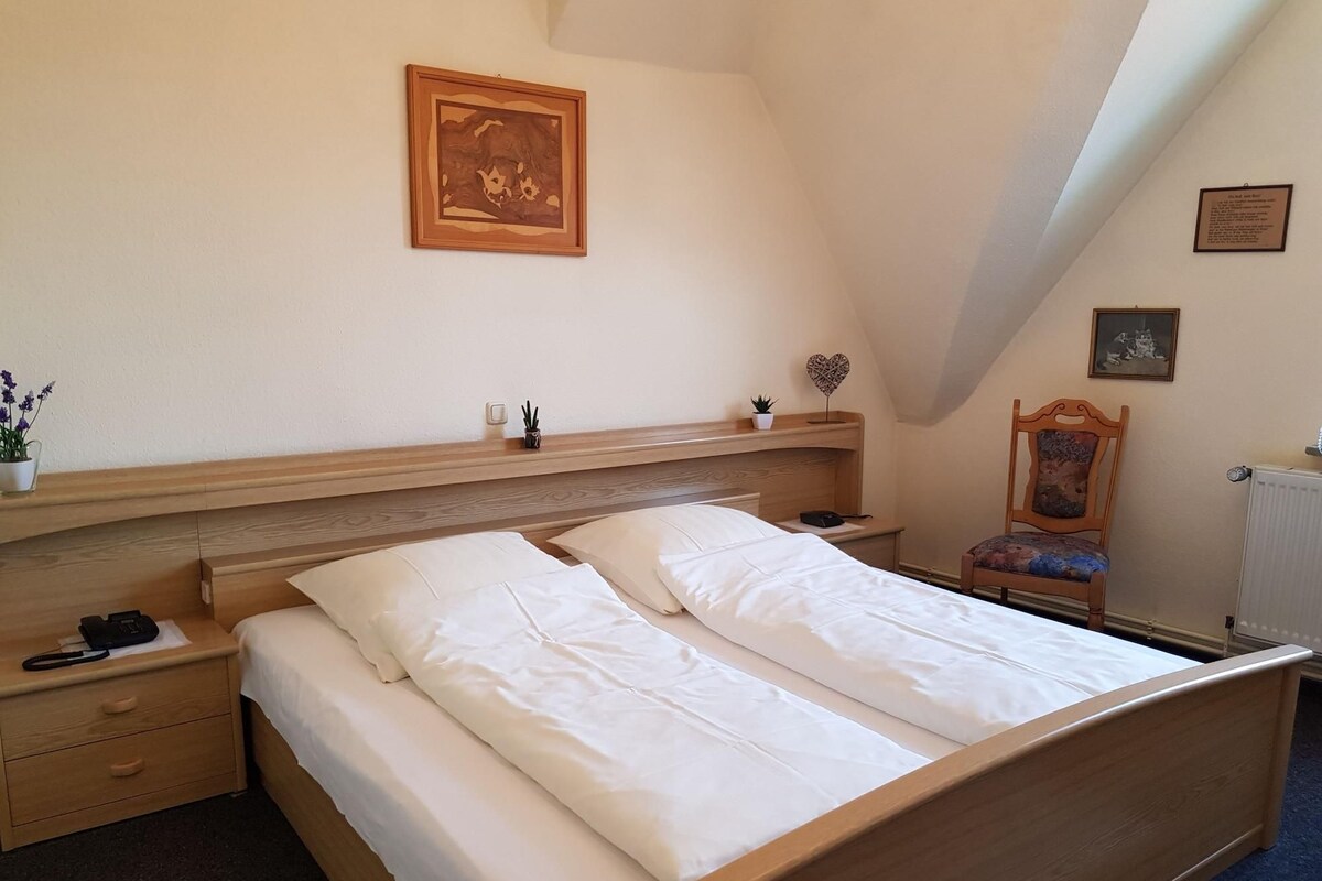 A bedroom with a wooden bed, a nightstand, a chair, and a small shelf with decorative items.