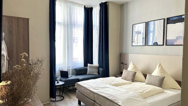 Moderni hotellihuone, jossa on suuri sänky, sohva, pieni pöytä ja työpöytä. Ikkunoissa on verhot, ja työpöydällä on maljakko, jossa on kuivattuja kukkia.