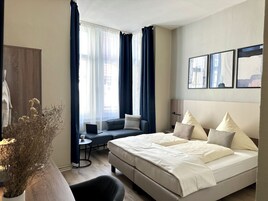 Ein modernes Hotelzimmer mit einem großen Bett, einer Couch, einem kleinen Tisch und einem Schreibtisch. An den Fenstern hängen Vorhänge, und auf dem Schreibtisch steht eine Vase mit Trockenblumen.