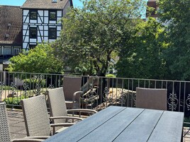 Eine Terrasse mit Korbmöbeln und einem Metallgeländer, mit Blick auf einen Baum und Gebäude im Hintergrund.
