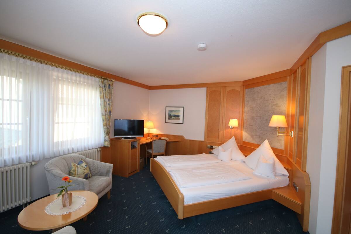 A hotel room with a bed, a desk, a chair, a TV, and a small table with a vase of flowers.