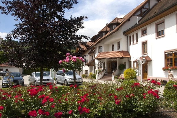 Un bâtiment traditionnel avec une façade blanche et un toit recouvert de tuiles, une entrée bien visible avec des marches, et des voitures garées au premier plan.
