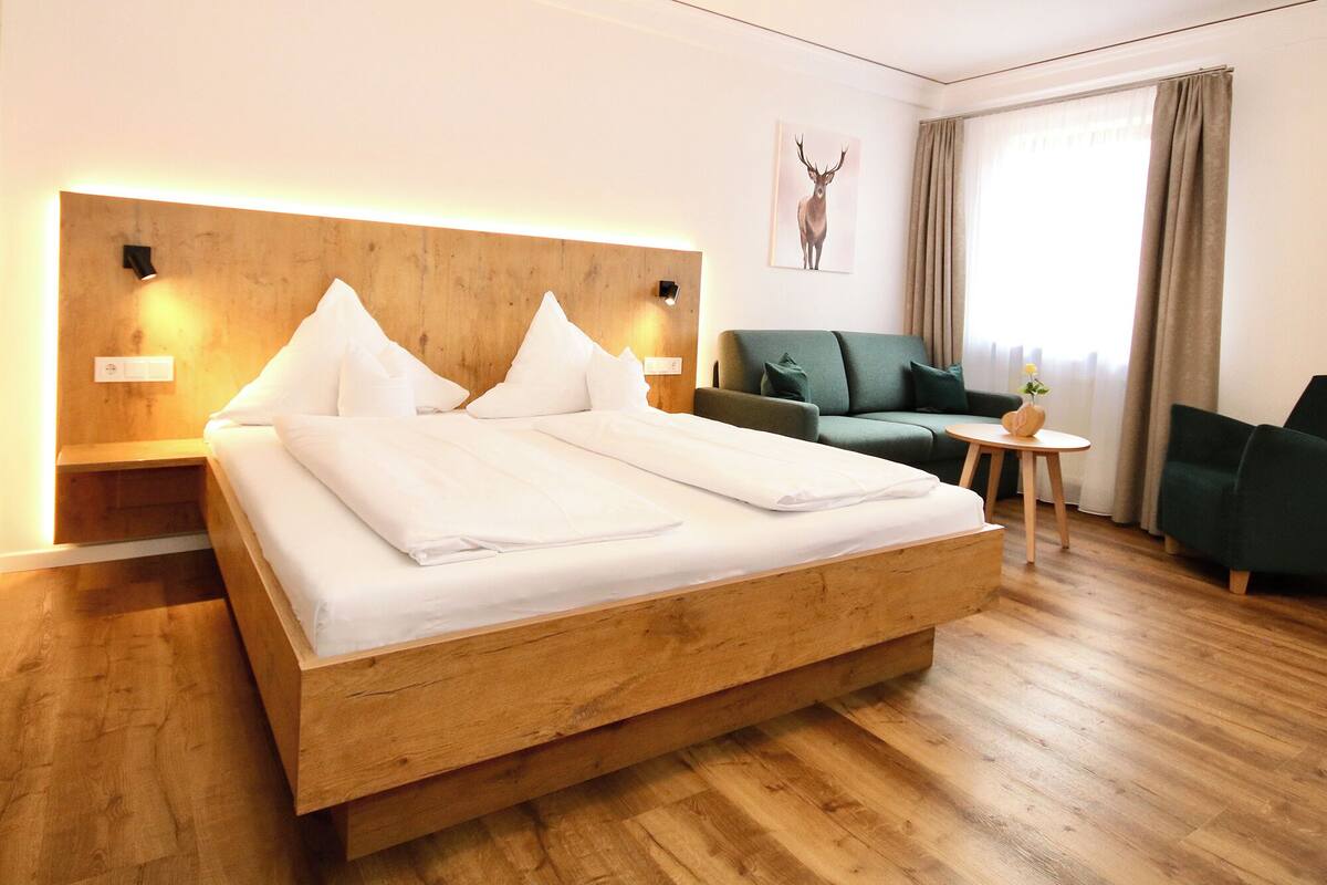 A hotel room with a large bed, a green sofa, a small table with a vase, and a deer artwork on the wall.