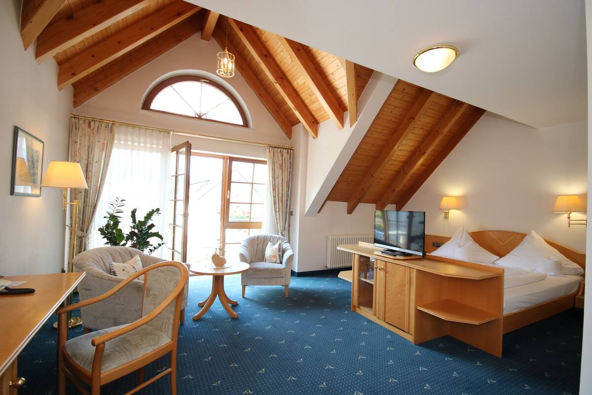A hotel room with a wooden ceiling, a bed, a desk, a chair, a TV, and a window with curtains.