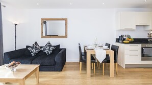 A modern living room with a dark grey sofa, a wooden coffee table, and a dining area with a wooden table and chairs. - Highlife Lexington Apartments (Slough)