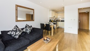 A modern living room with a dark grey sofa, a wooden coffee table with a bowl of fruit, and a large mirror on the wall. - Highlife Lexington Apartments (Slough)