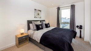 A modern bedroom with a large bed, a nightstand with a lamp, a decorative wall piece, and a view of the city through a window. - Highlife Lexington Apartments (Slough)