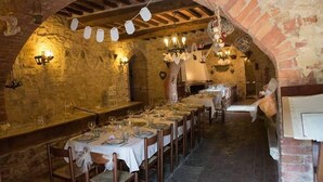 A long dining table set with white tablecloths and chairs, in a rustic stone-walled room with exposed wooden beams and hanging light fixtures. - Verdidea - Aia Vecchia (Asciano)