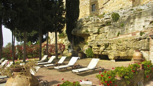 A stone building with a rock wall, outdoor seating with sun loungers, and potted plants with red flowers. - Verdidea - Aia Vecchia (Asciano)
