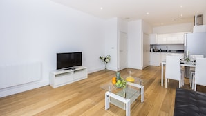A modern living room with a flat-screen TV, a glass coffee table with fruit, a sofa, and a dining area with white chairs.