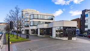 A modern building with a glass facade, a small outdoor seating area, and a visible entrance.