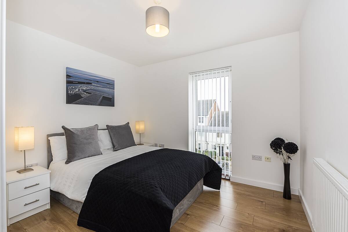 A bedroom with a bed, bedside tables, a nightstand, a lamp, a painting, a window with blinds, and a vase with flowers.