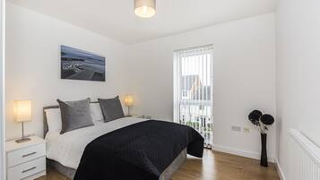 A bedroom with a bed, bedside tables, a nightstand, a lamp, a painting, a window with blinds, and a vase with flowers.