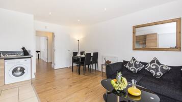 A modern living room with a black sofa, a dining area with a table and chairs, and a kitchen with a washing machine.
