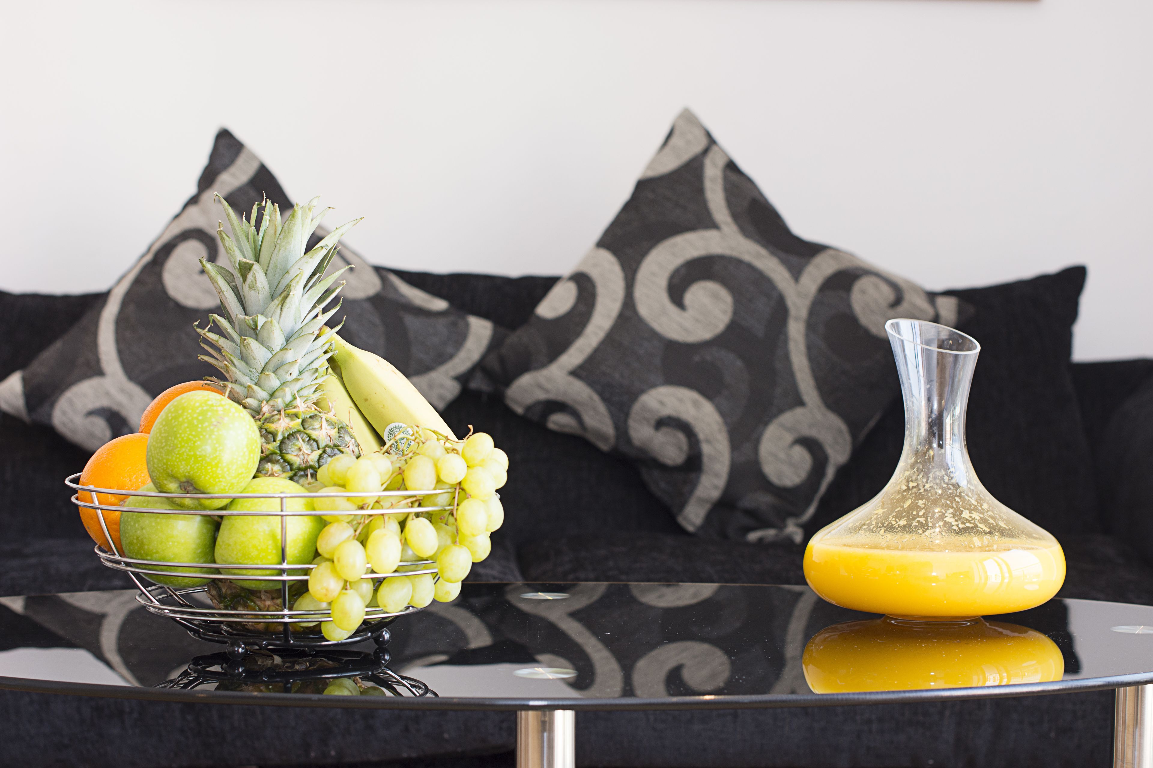 Un frutero con manzanas, uvas y una piña sobre una mesa reflectante, junto a un vaso de jugo de naranja.