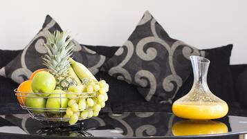 Un frutero con manzanas, uvas y una piña sobre una mesa reflectante, junto a un vaso de jugo de naranja.