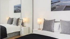 A modern hotel room with a large mirror, a bed with grey bedding, bedside tables with lamps, and a framed picture on the wall. - Merrick House Apartments (Reading)