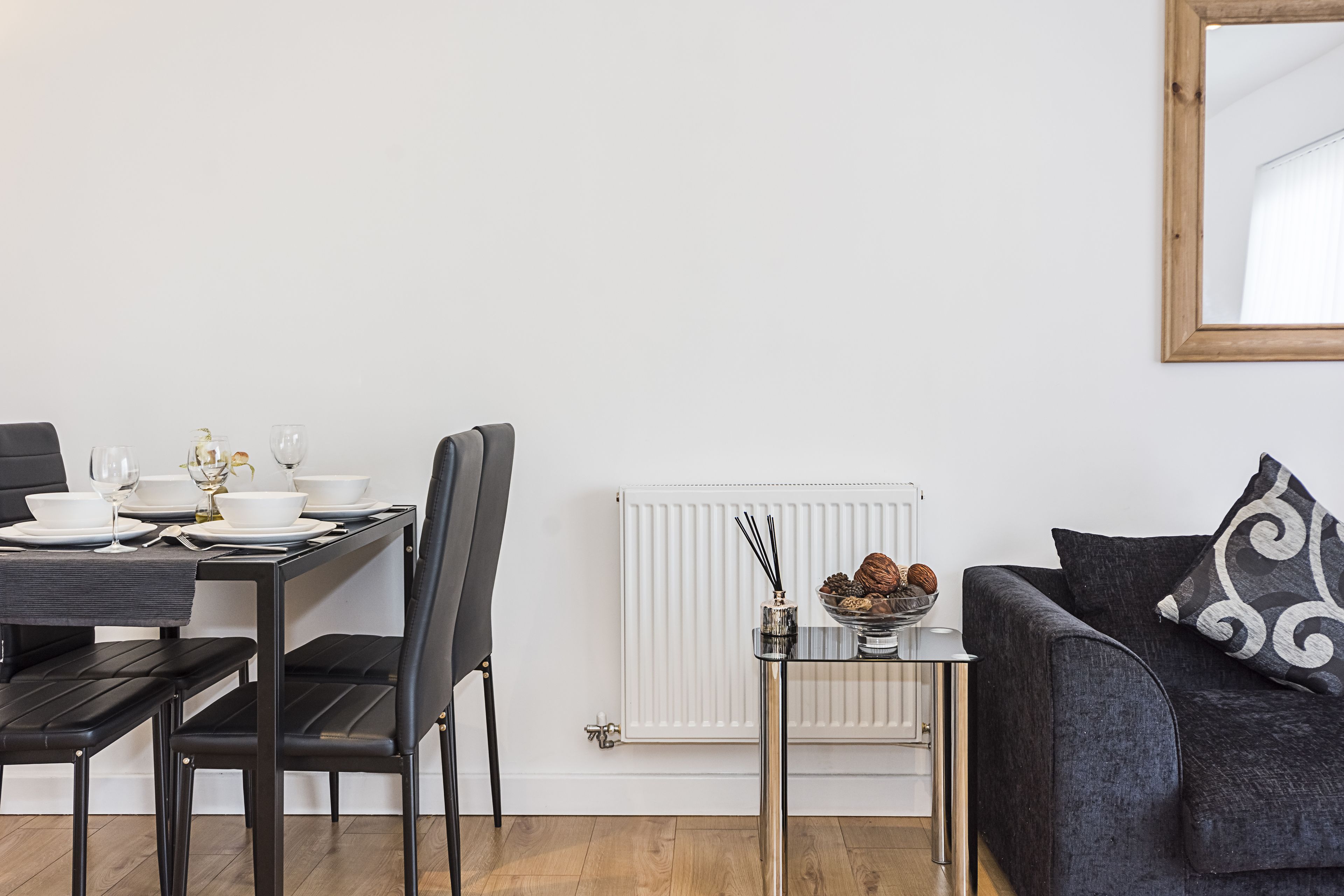 Ein modernes Wohnzimmer mit Essbereich. Es umfasst einen schwarzen Esstisch mit weißen Tellern und Gläsern, ein schwarzes Sofa mit einem gemusterten Kissen und einen kleinen Glastisch mit einer dekorativen Schüssel.