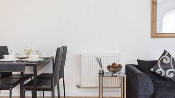 Ein modernes Wohnzimmer mit Essbereich. Es umfasst einen schwarzen Esstisch mit weißen Tellern und Gläsern, ein schwarzes Sofa mit einem gemusterten Kissen und einen kleinen Glastisch mit einer dekorativen Schüssel.