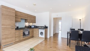 A modern kitchen with wooden cabinets, a built-in oven, and a dishwasher. There is a dining area with a table and chairs. - Merrick House Apartments (Reading)