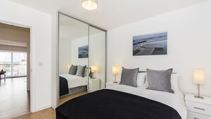 A bedroom with a large bed, a sliding door to a balcony, and a painting of a coastal scene. - Merrick House Apartments (Reading)