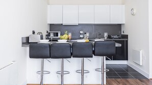 A modern kitchen with white cabinets, a black countertop, and four black bar stools. - Heron House Apartments (Reading)