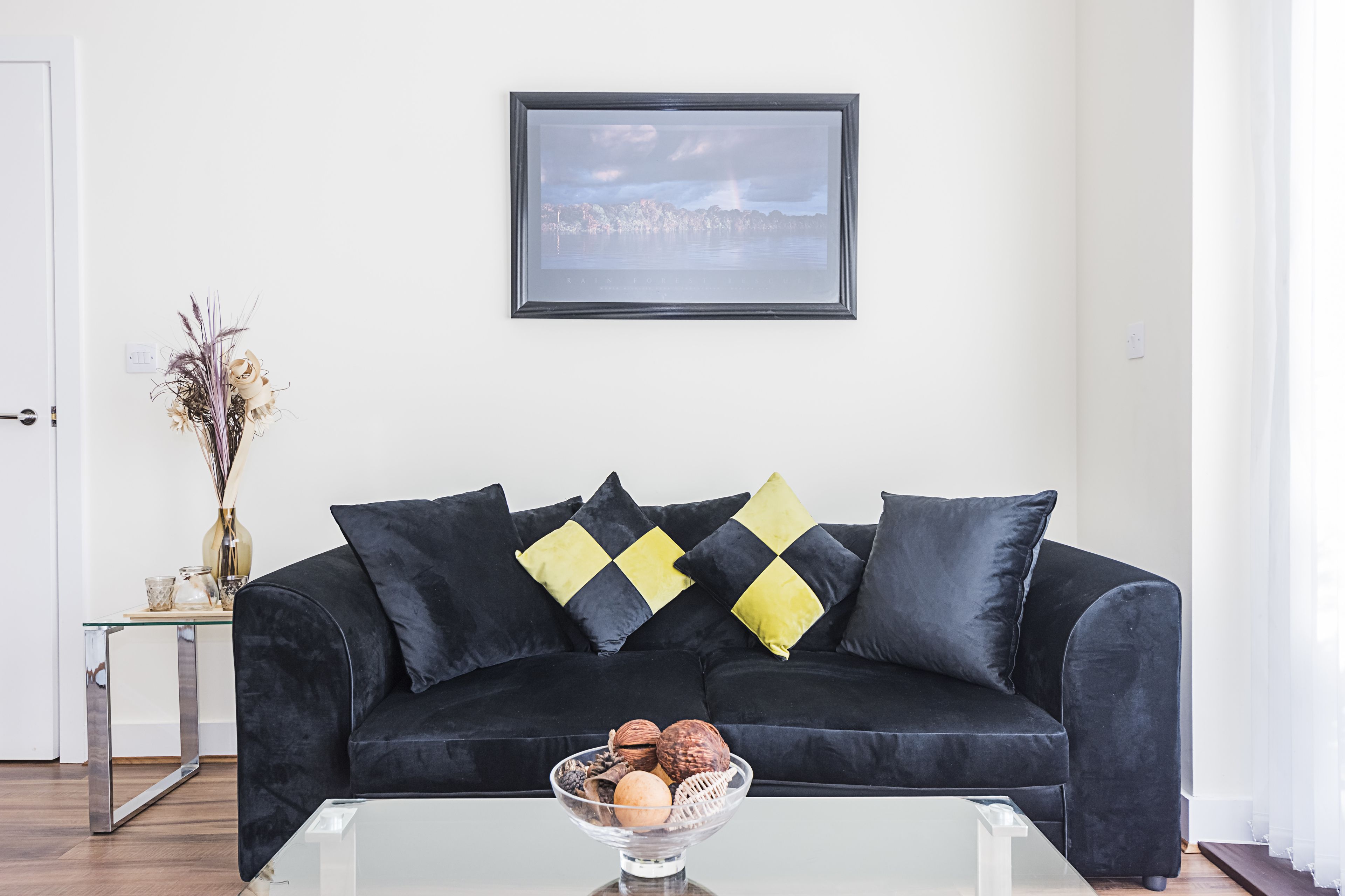 Un salon moderne avec un canapé noir, une table basse en verre et un tableau encadré accroché au mur.
