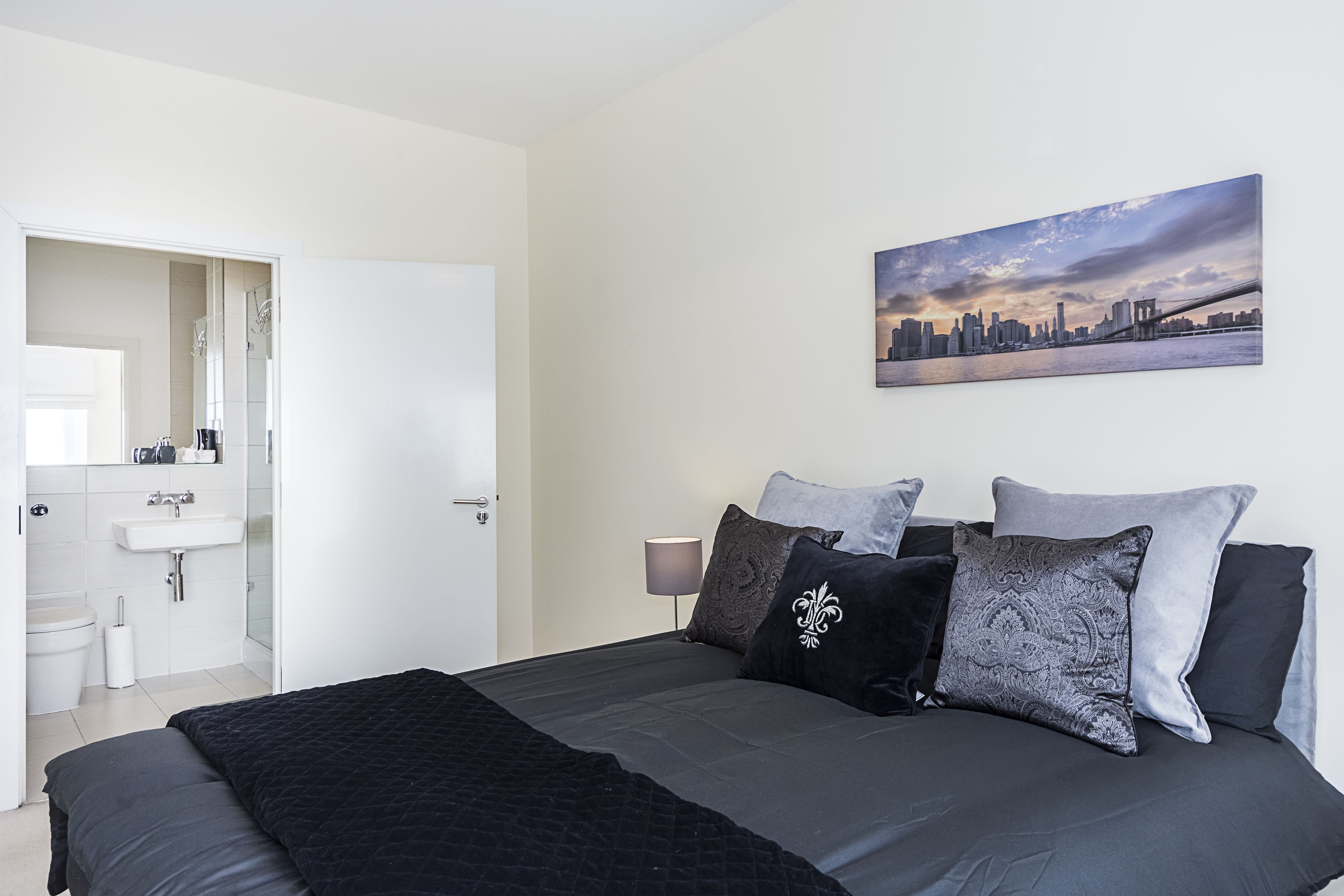 A bedroom with a large bed, a cityscape painting, and a bathroom visible through an open door.