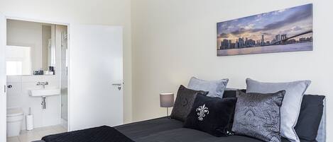 A bedroom with a large bed, a cityscape painting, and a bathroom visible through an open door.