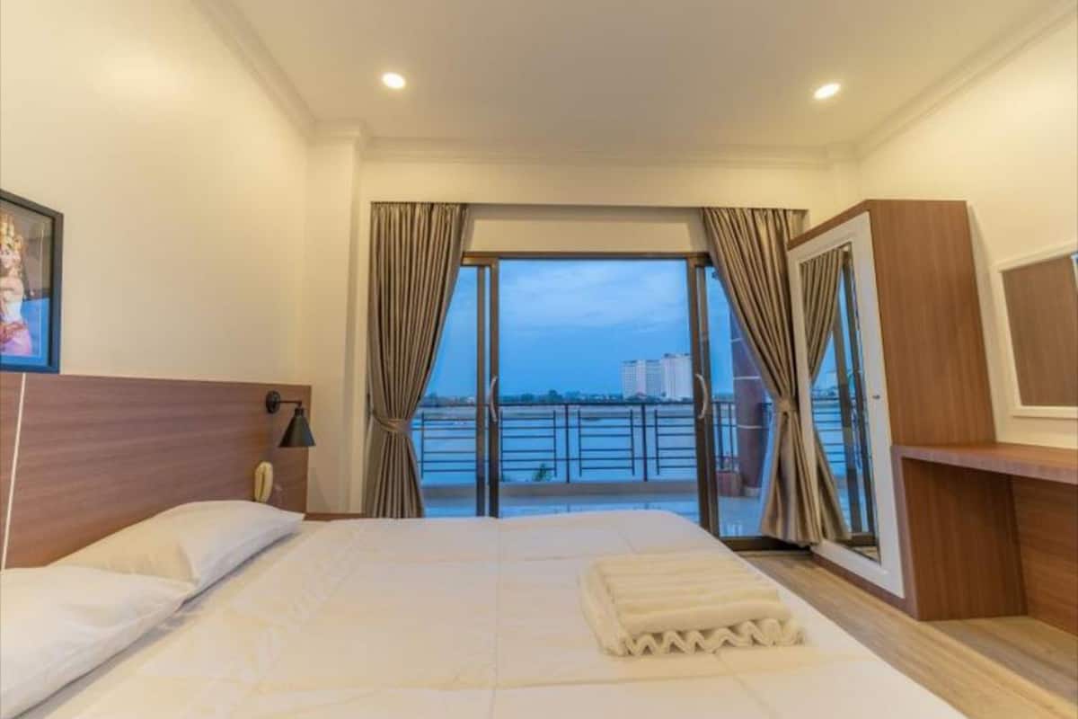 A hotel room with a large bed, a wooden headboard, a bedside table with a lamp, a TV, and a view of a body of water and buildings.