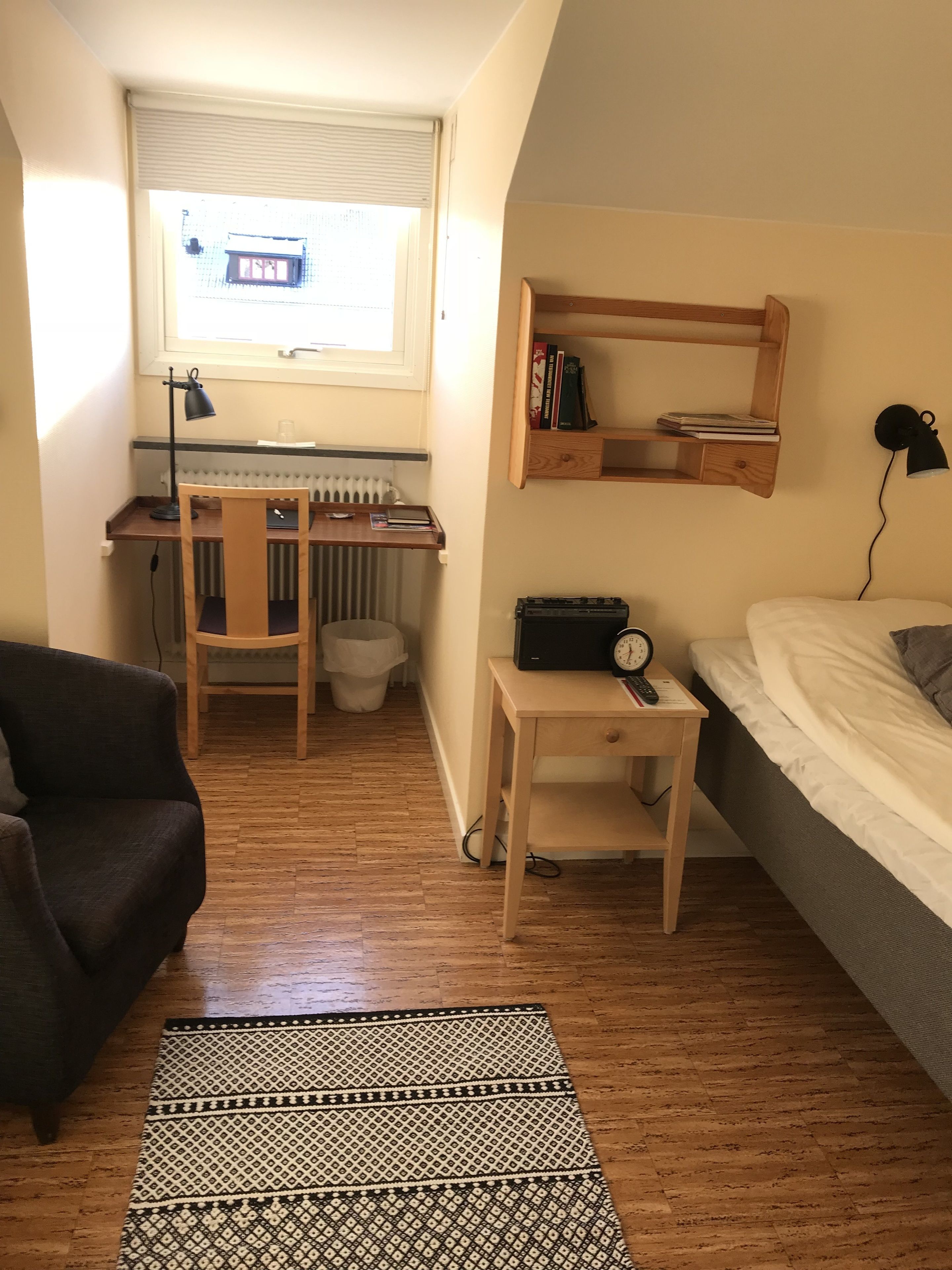 Una habitación compacta con escritorio, silla, cama y una mesita con un reloj.