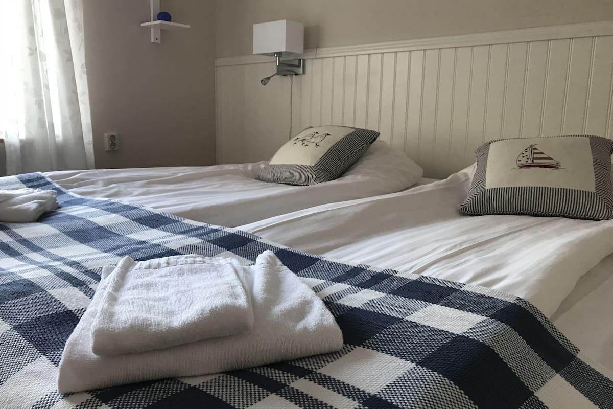 A hotel room with two beds, each with a checkered bedspread and pillows with a bird design. There is a white wall-mounted light above the beds and a white shelf with a blue object on it.