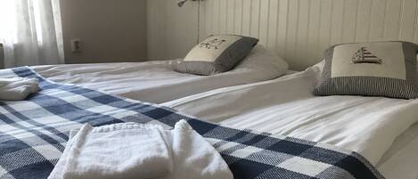 Une chambre d’hôtel avec deux lits, chacun recouvert d’une couvre-lit à carreaux et agrémenté d’oreillers décorés de motifs d’oiseaux. Au-dessus des lits, un luminaire blanc est fixé au mur, et une étagère blanche contient un objet bleu.