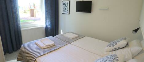 Cama doble con cubrecama a rayas y dos almohadas decorativas, un televisor de pantalla plana montado en la pared y una ventana con cortinas.