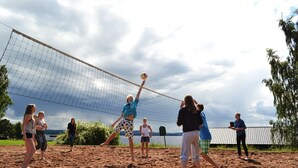 A group of people playing volleyball on a sandy court with a net, surrounded by trees and a cloudy sky.