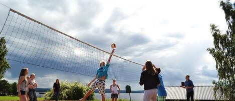 Un grupo de personas jugando voleibol en una cancha de arena, rodeado de árboles y un cielo nublado.
