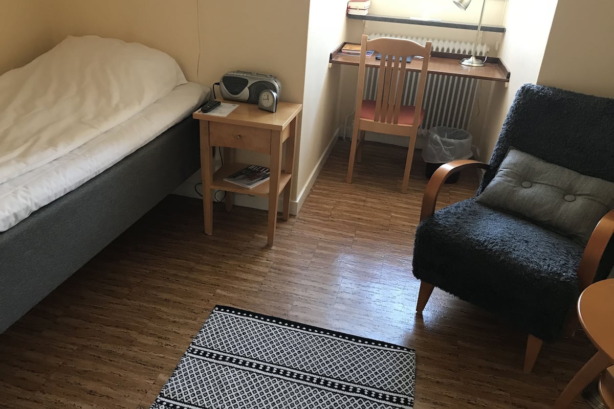 A small, cozy room with a bed, a desk, a chair, and a patterned rug.