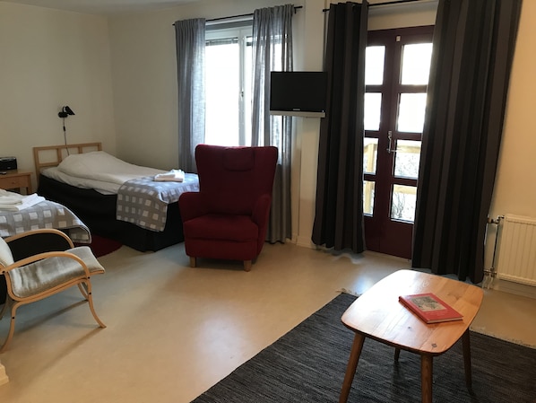 Quarto de hotel com duas camas, uma poltrona vermelha, uma mesa de madeira e uma televisão.