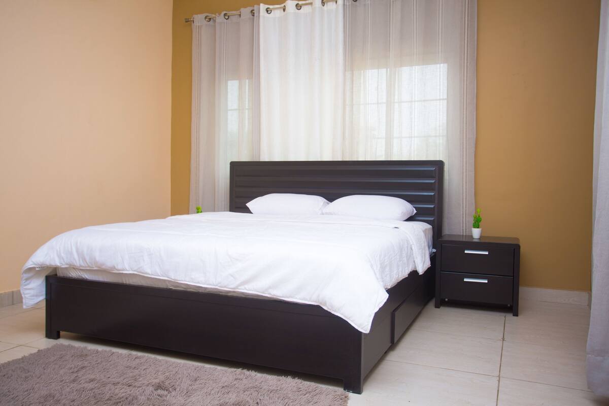 A bedroom with a dark wooden bed, white bedding, a small bedside table with drawers, and sheer curtains.