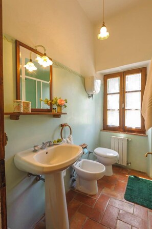 Een badkamer met een wastafel op een voetstuk, twee toiletten, een spiegel, een raam met een handdoek en een radiator.