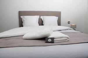 Una cama bien hecha con sábanas blancas y una toalla doblada, una tarjeta negra pequeña sobre la cama, y una lámpara de noche.